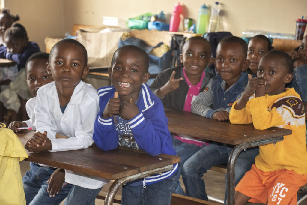 Young students at school desks smile and give thumbs-up.